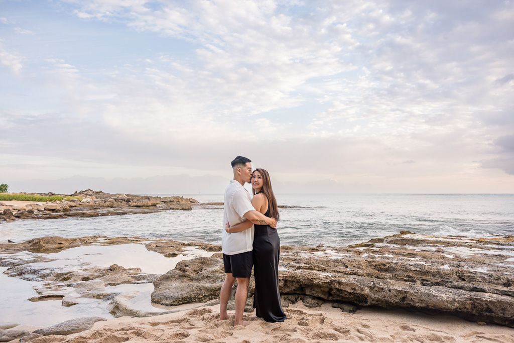 Groom-to-be wraps his arms around his fiancée as they stand on the rocky shore of Ko Olina lagoon in Oahu, gazing at the sunset over the water.