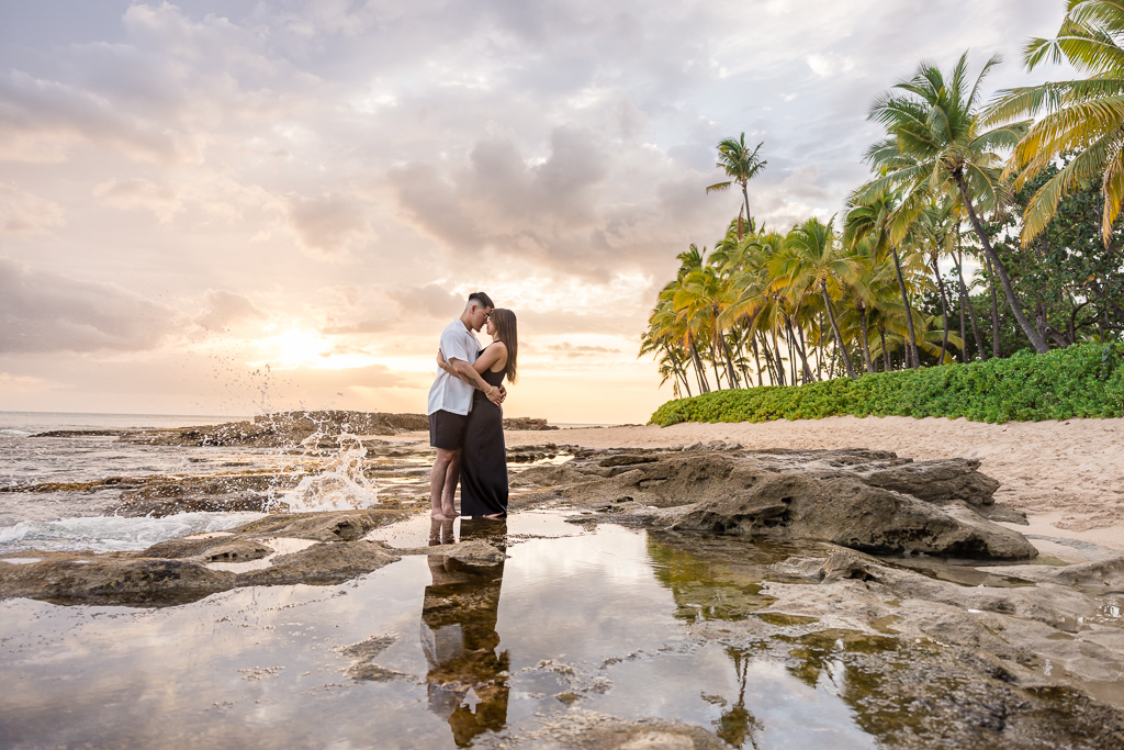 Groom in white and bride in black dress embraces for an engagement shoot on Oahu's Ko Olina lagoon beach at sunset with palm trees and ocean.