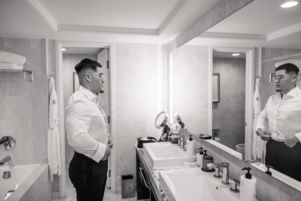 black-and-white photo of groom buttoning up his shirt in front of a bathroom mirror