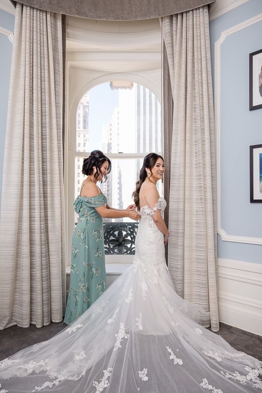 sister helping bride button up her dress in front of a window