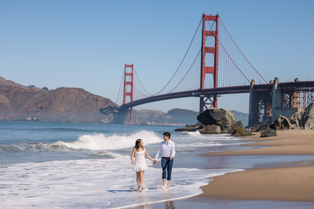 engagement photo of couple running through the water at the beach in front of the Golden Gate Bridge