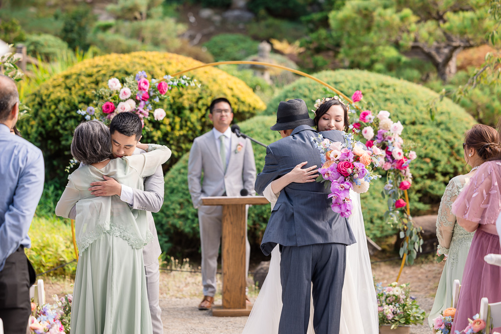 the bride and groom hugging the bride’s parents at the end of the aisle at the hand-off