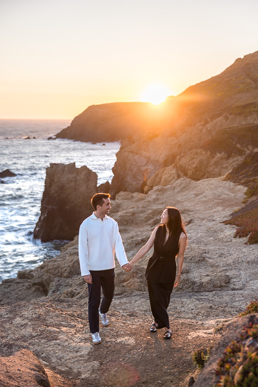 couple walking along coastal ocean bluff rocks with the sun setting behind them