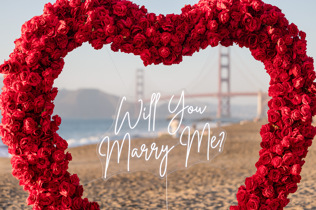 Will Your Marry Me neon lit sign hanging inside floral heart on the beach