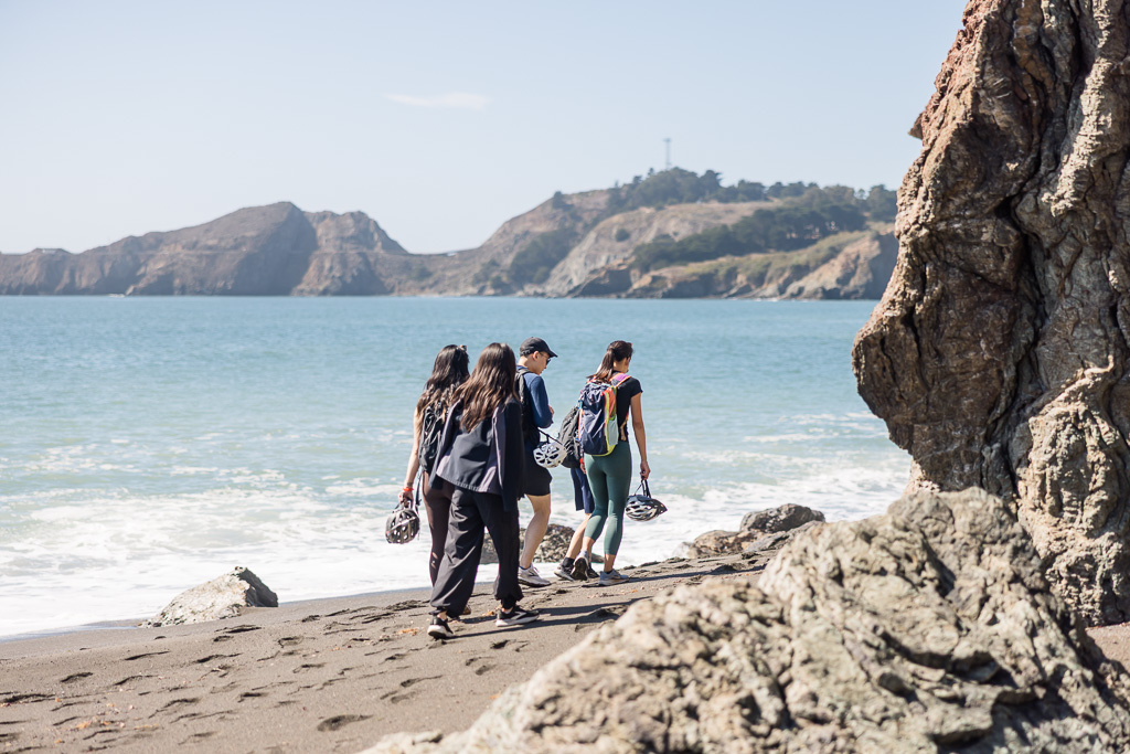 sneaky photo of a group of friends walking along the beach with bike helmets in hand