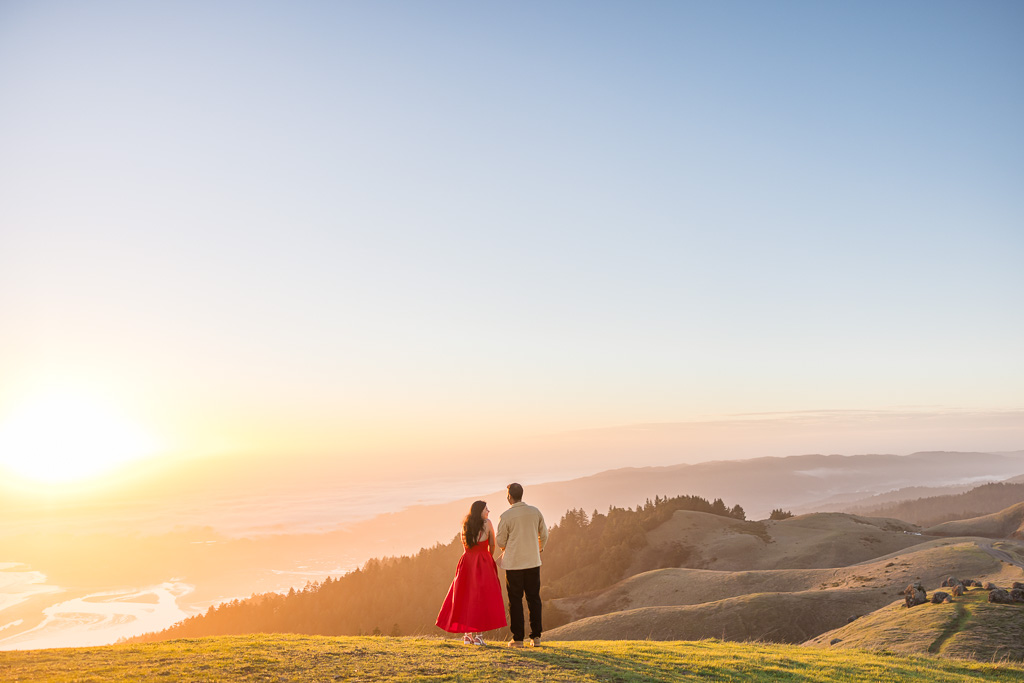 couple in tan outfit and red dress standing atop mountain at golden hour looking off into the hills and ocean
