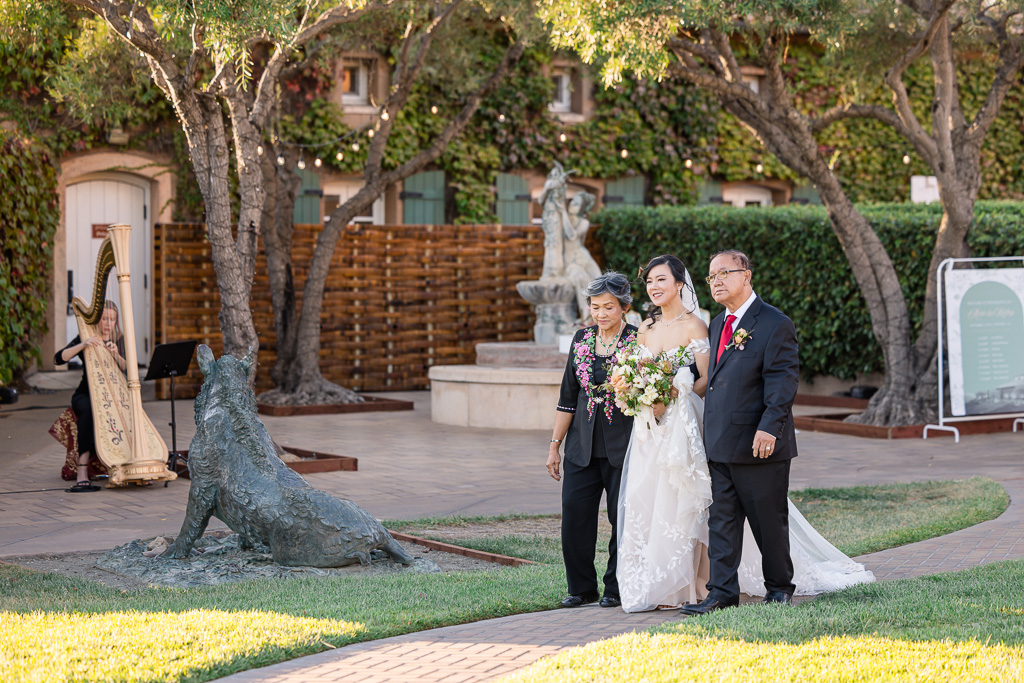 the bride walking down the aisle with her parents on either side and a harpist in the background