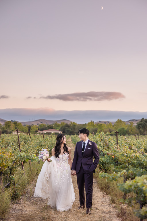 newlyweds walking along vineyards at dusk with pink and purple sky above them