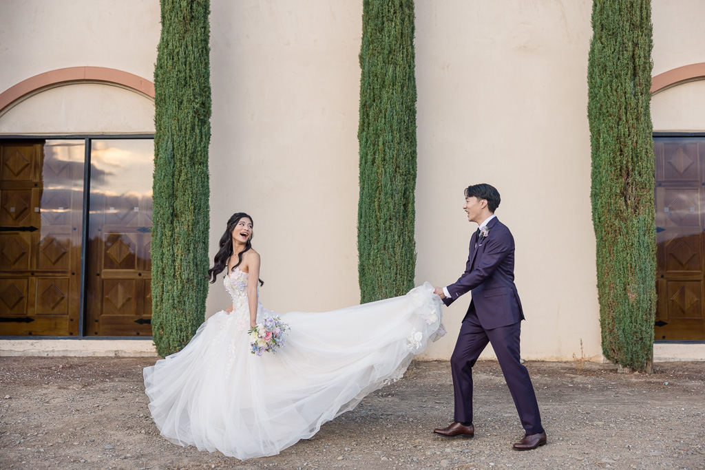 Groom holding bride's wedding dress train outside venue