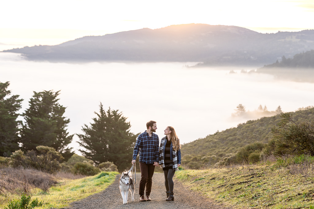 engagement photos of a couple walking their dog on a hiking trail with mountains and a sea of fog in the background