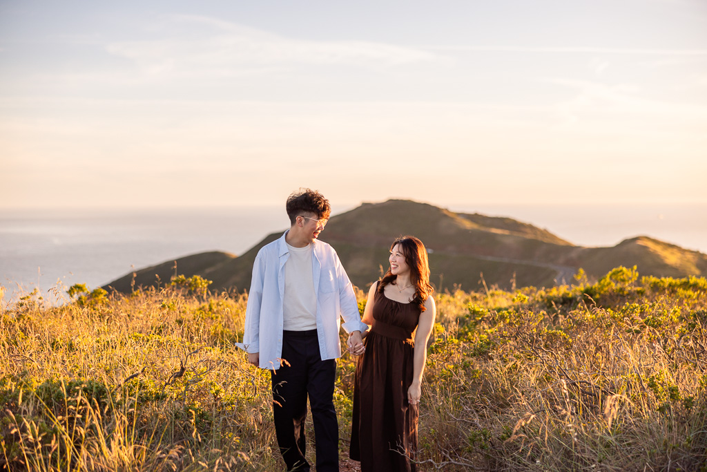 couple walking through golden field of grass, holding hands, at sunset hour