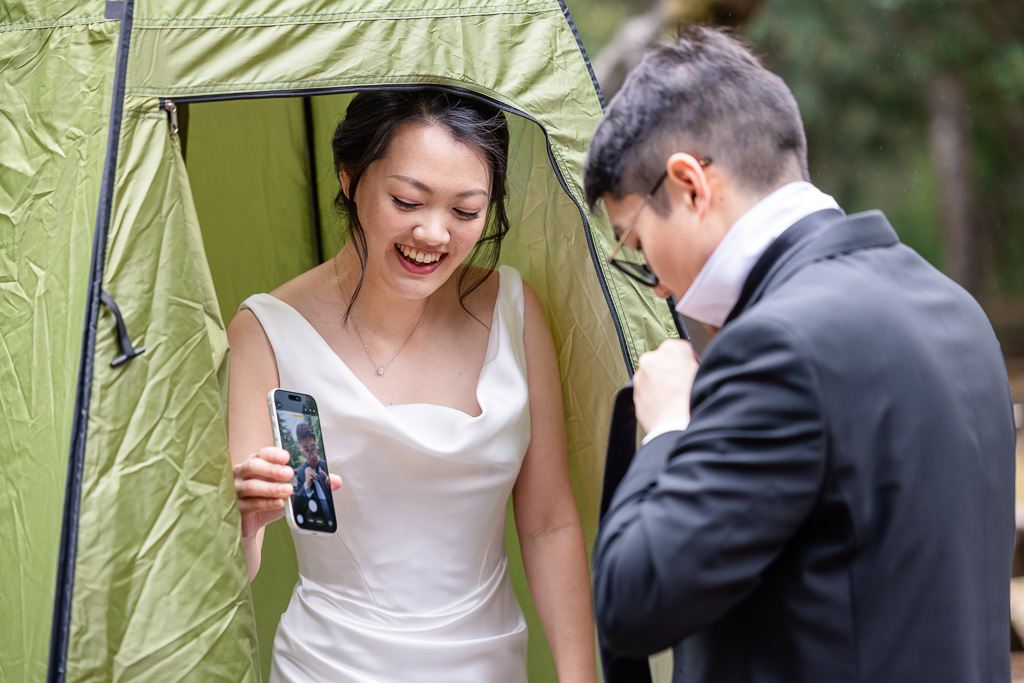 bride holding up a phone camera to help groom see his tie