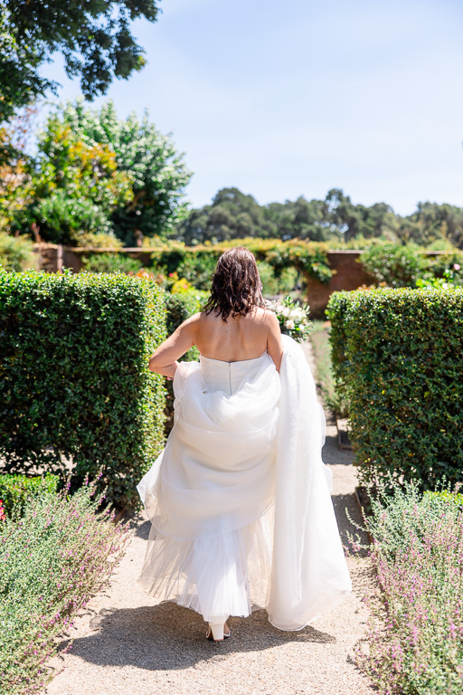 candid back shot of bride holding up her owno dress and walking through a garden by herself