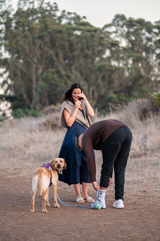 girl starting to wipe away a tear while her boyfriend bends down to pull ring box out of pant leg