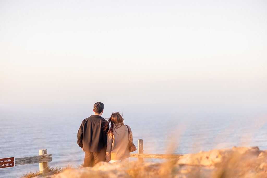 Photographer hidden behind tall grass capturing couple standing together at Point Reyes South Beach Overlook, facing the ocean in the soft evening light.