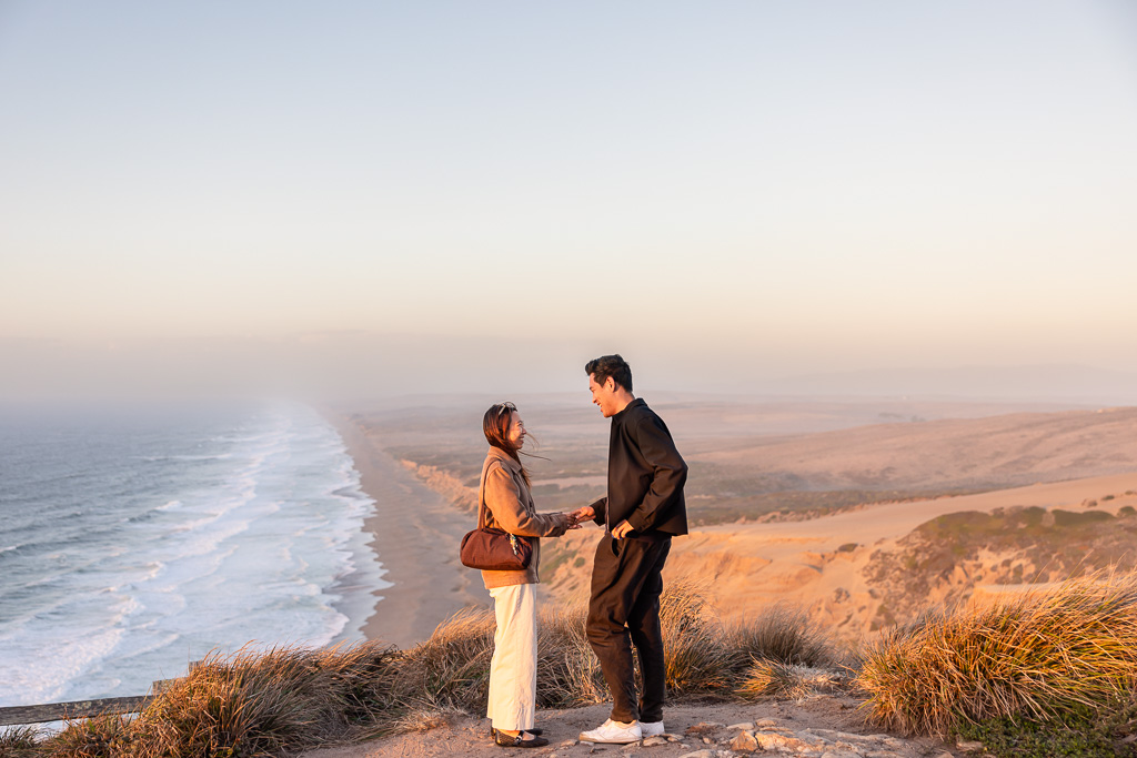 At the coastal overlook, a man and woman hold hands and smile at each other above the long shoreline just before proposal moment