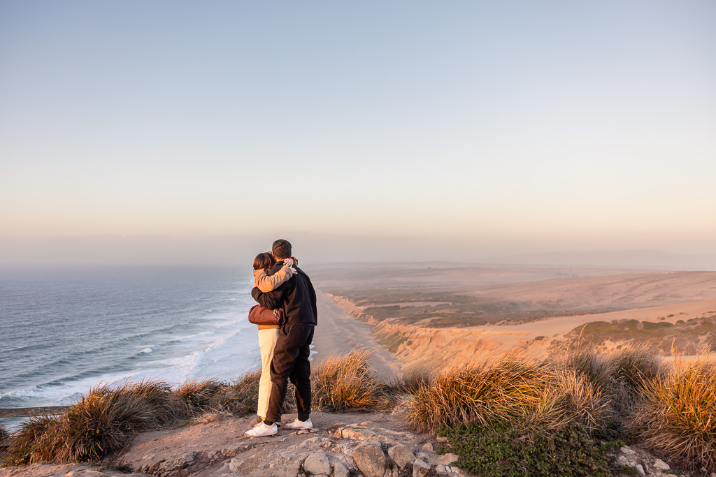 The couple embraces after the proposal, with the rugged Point Reyes coast stretching behind them.