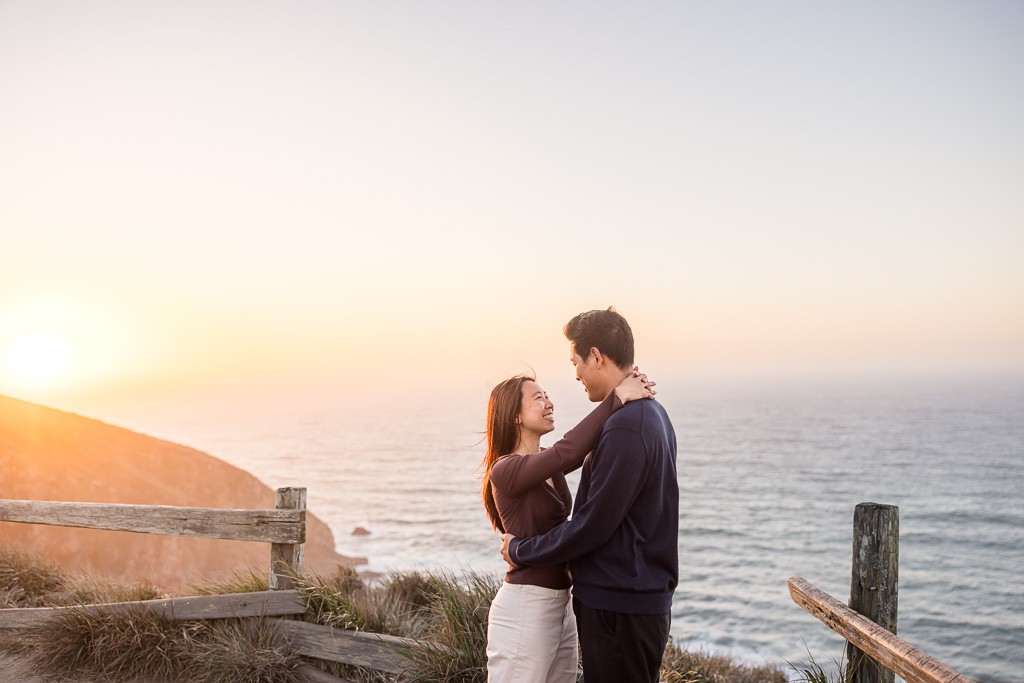 A couple embraces beside the weathered overlook fence as the sun sets over the ocean during golden hour