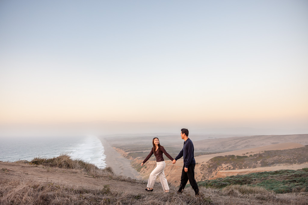Wide view of the couple holding hands along the grassy bluff above the Point Reyes Beach