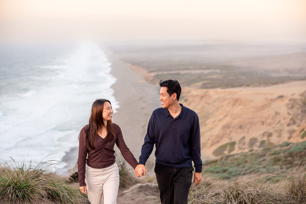 The couple walk hand in hand along the bluff during their Point Reyes engagement portraits over the beach behind them
