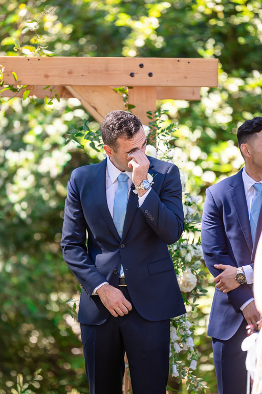 Groom wipes away a tear beneath the floral ceremony arch as the bride enters