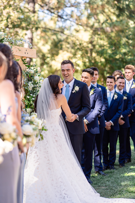 Bride and groom face each other beneath the floral arch with groomsmen behind them