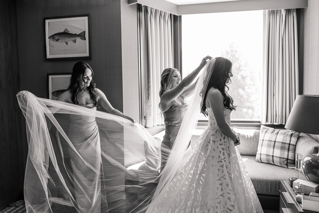 Black-and-white wedding portrait of the bride in her dress beside the window as bridesmaids help with her veil