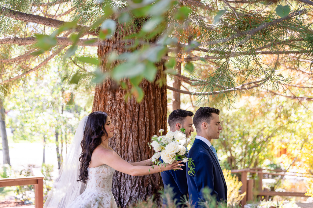 Bride holds her bouquet behind her brothers for their first look beneath a pine tree