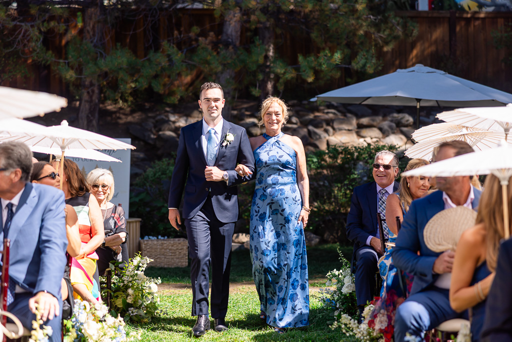 Groom walks up the ceremony aisle with his mom as attendees wait beneath parasols