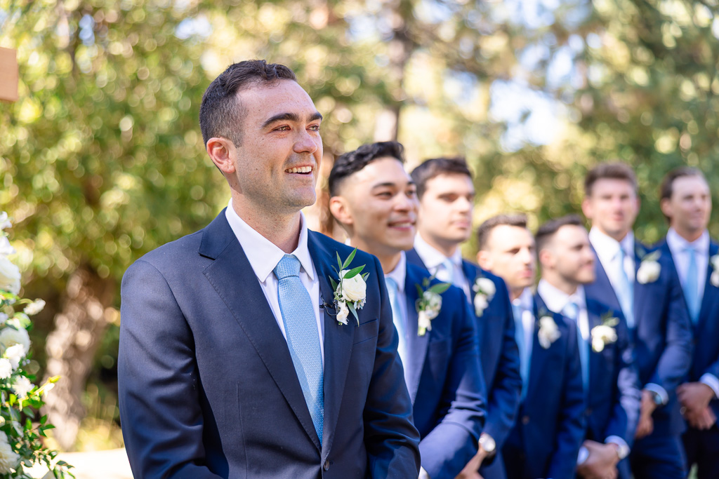 Groom waits beneath the trees with his groomsmen lined up behind him
