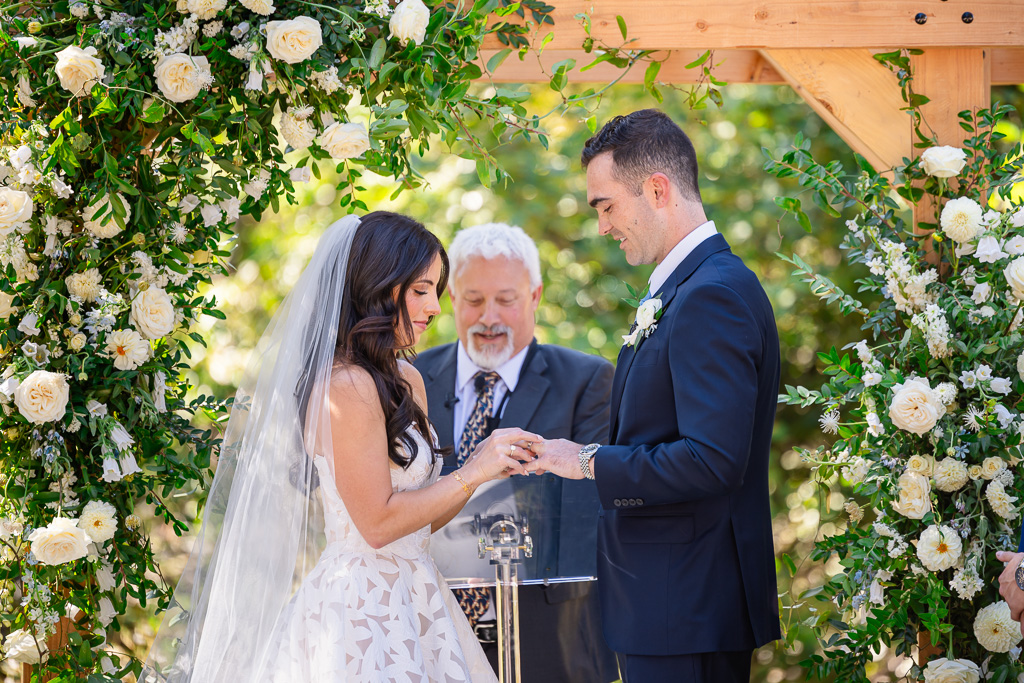 Bride and groom exchange wedding rings beneath the floral arch
