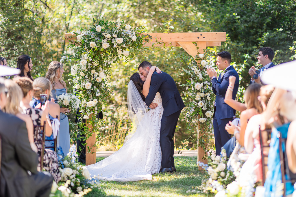 Bride and groom kiss beneath the floral arch at their wedding on the Creekside Lawn