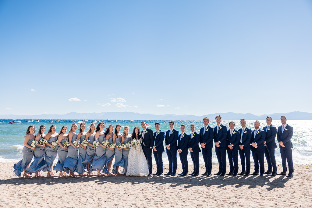 Bride, groom, and bridal party lined up neatly for a photo on the beach of Lake Tahoe under the afternoon sun