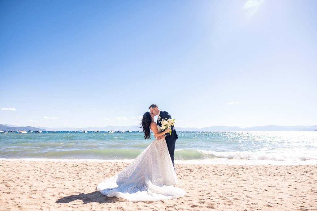 Bride and groom kiss on a Lake Tahoe beach against deep blue water and a clear sky