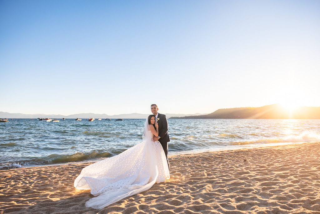 Bride and groom pose together on the Lake Tahoe beach at sunset, showing off long dress
