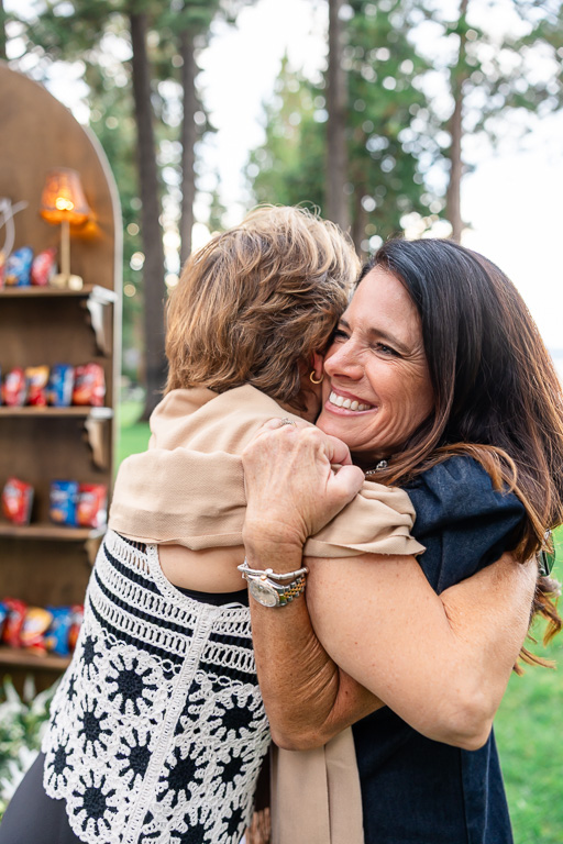 Mother of the bride embraces a guest beneath the pines during the couple’s pre-wedding gathering.
