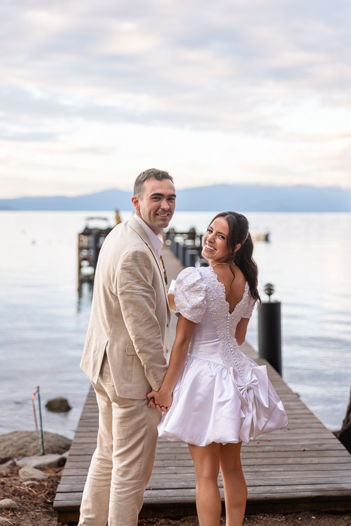 The bride and groom look back at the camera while holding hands on a dock above the water of Lake Tahoe