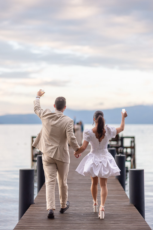 The bride and groom walk hand in hand down the dock toward the lake, raising their arms holding drinks