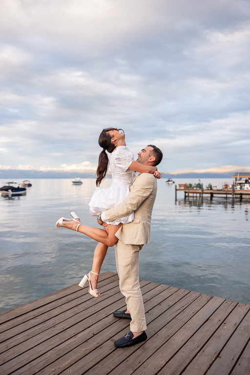 The groom lifts the bride on a dock as mountains and boats dot the lake behind them.