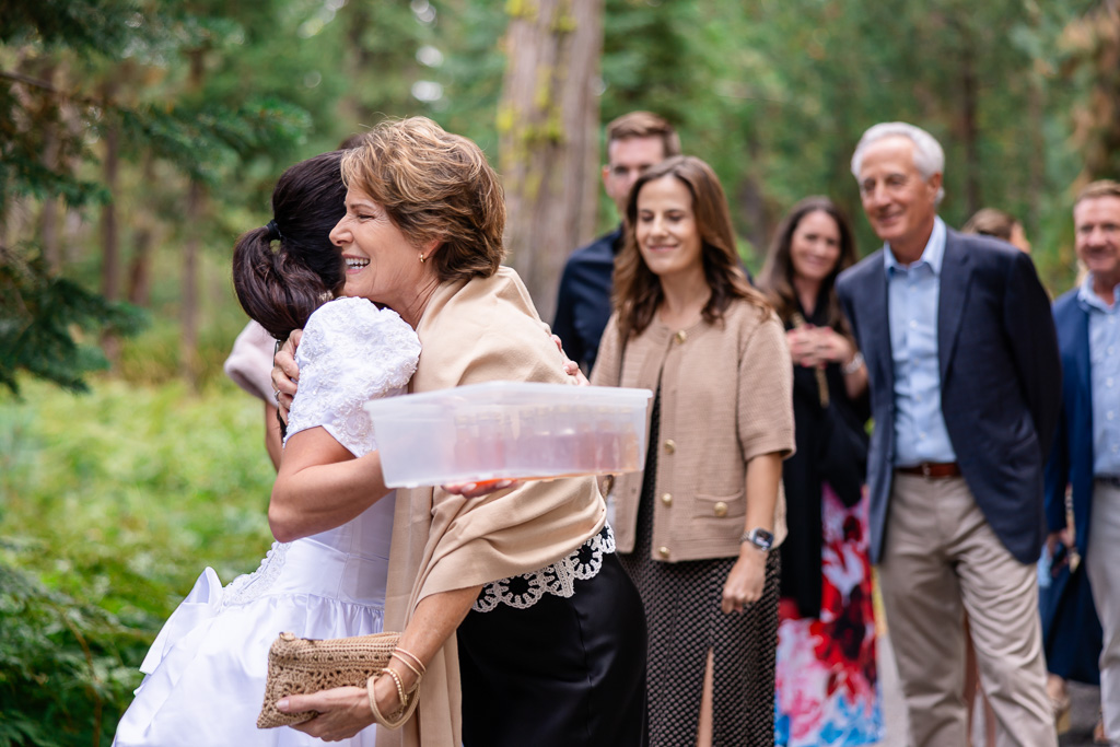 The bride embraces a guest near the driveway beneath the pines.