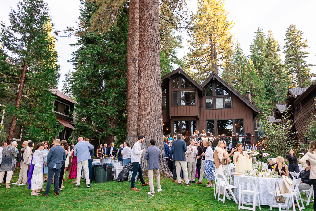 Friends mingle on the lawn beside a pine-lined lakeside home during pre-wedding welcome reception