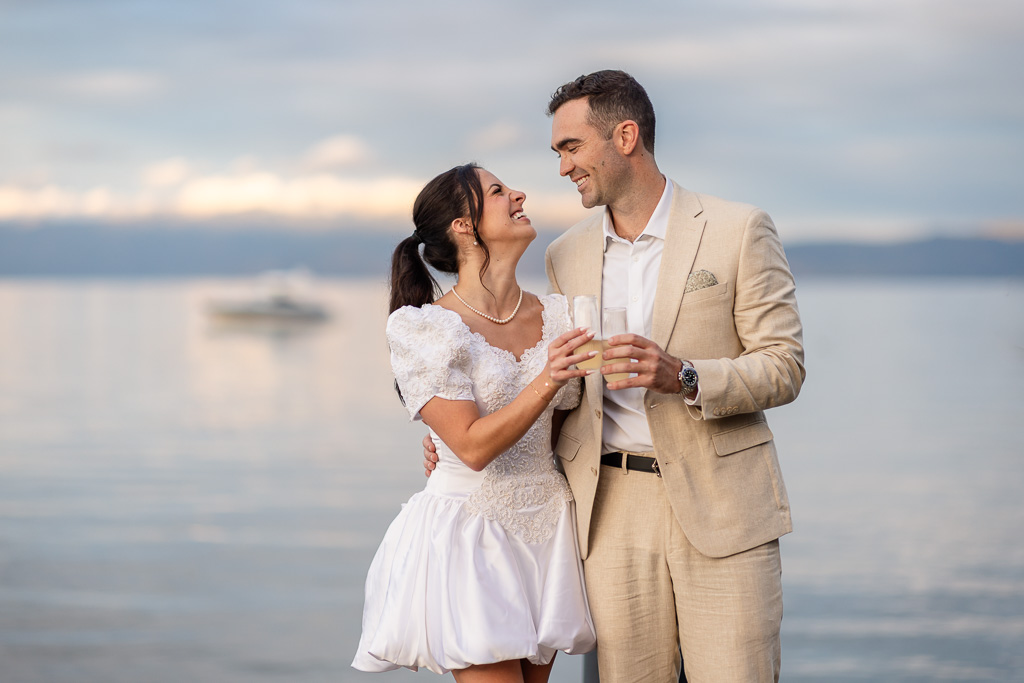 The bride and groom smile together over champagne near the lake.