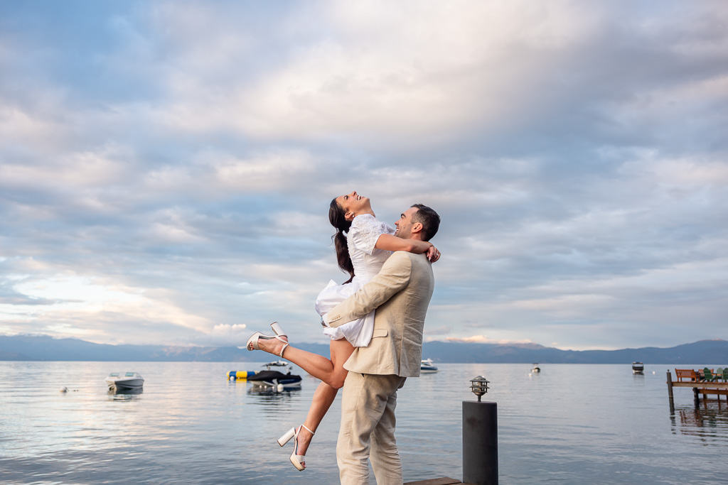 The groom lifts his laughing bride above the dock, with Lake Tahoe behind them.
