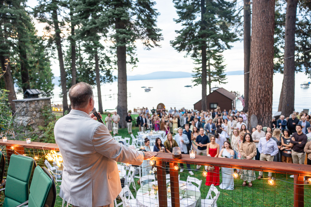 Father of the groom gives a heartfelt speech from the deck as guests gather below among the trees.
