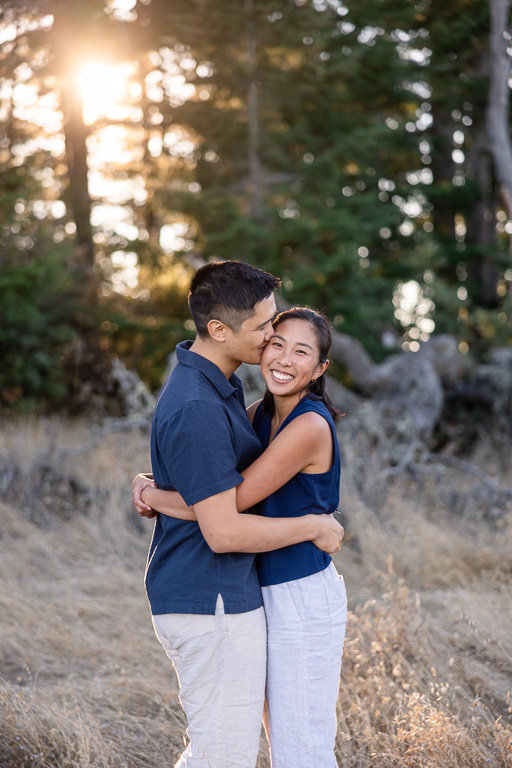 A man kisses his smiling fiancee on the cheek among the rocky, tree-lined trails of Mount Tamalpais.