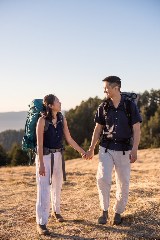 A couple in hiking packs hold hands and smile at one another on a sunlit Mount Tamalpais ridge.