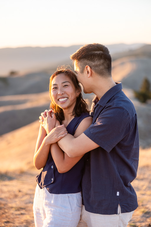 Engagement photo with man embracing her from behind in golden hillside light.