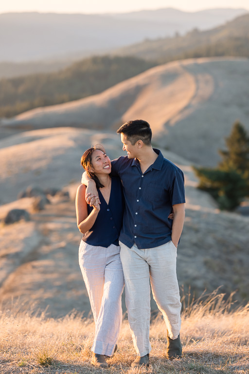 A couple walks arm in arm on a grassy hill, sharing a smile during their Mount Tamalpais engagement portraits.