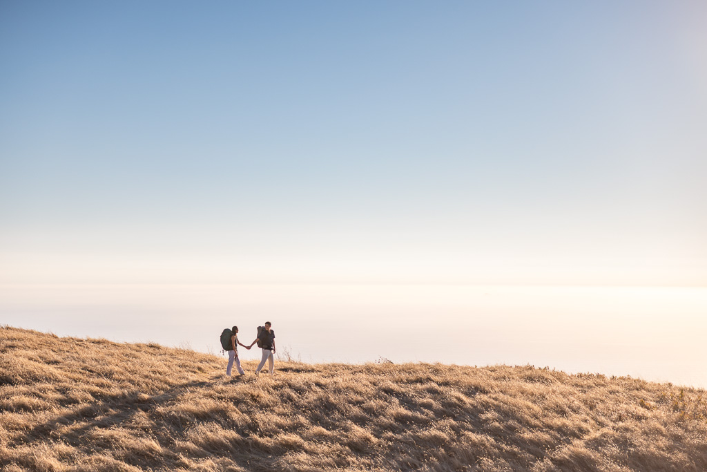 Wide sunset view of a couple hiking hand in hand across an open Mount Tamalpais ridgeline.