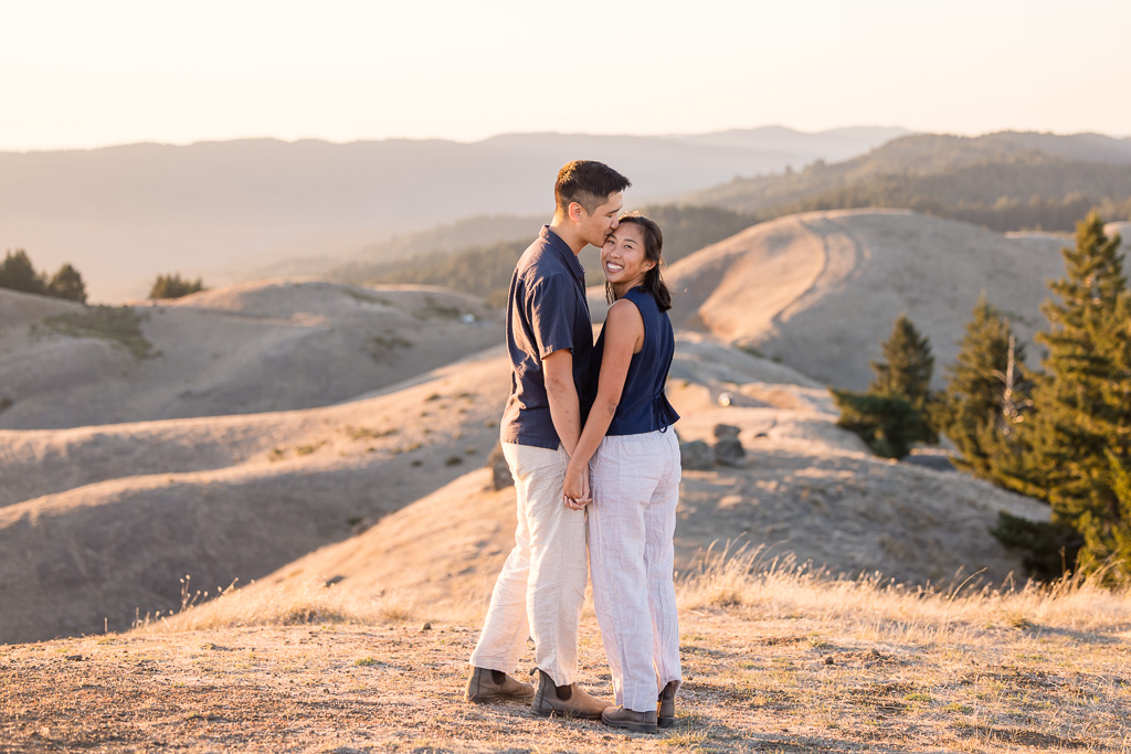 A man kisses his partner while she turns toward the camera in a warm, close engagement portrait.